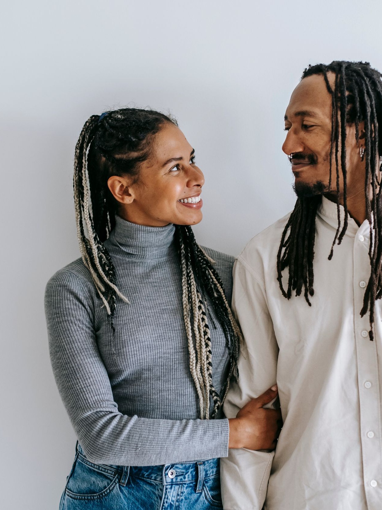 A couple with braids smiling at each other against a light wall
